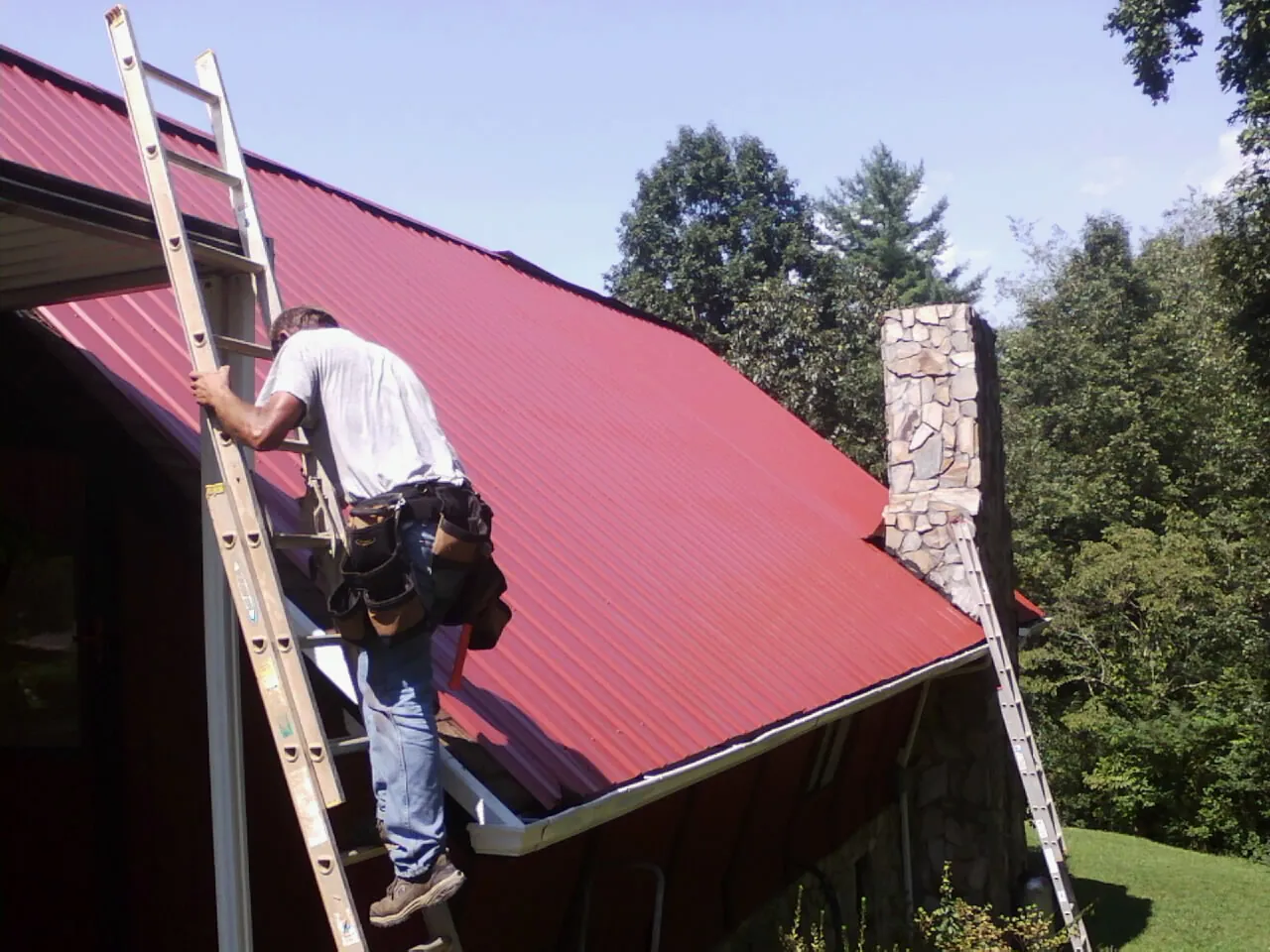 Roofer on a ladder beside a red metal roof and stone chimney — Soffit & Fascia Repair by Roofing Brothers in Brazil